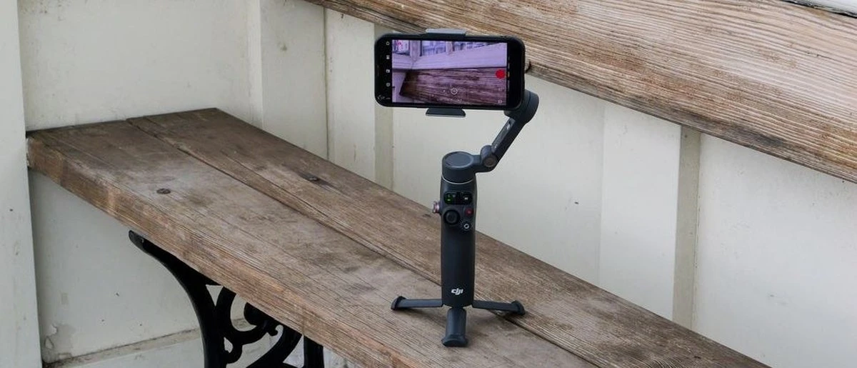 Стабілізатор DJI Osmo Mobile 7P стоїть на вбудованій підставці й тримає смартфон на дерев'яній стійці